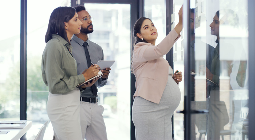 pregnant woman points at a white board describing work to her two colleagues