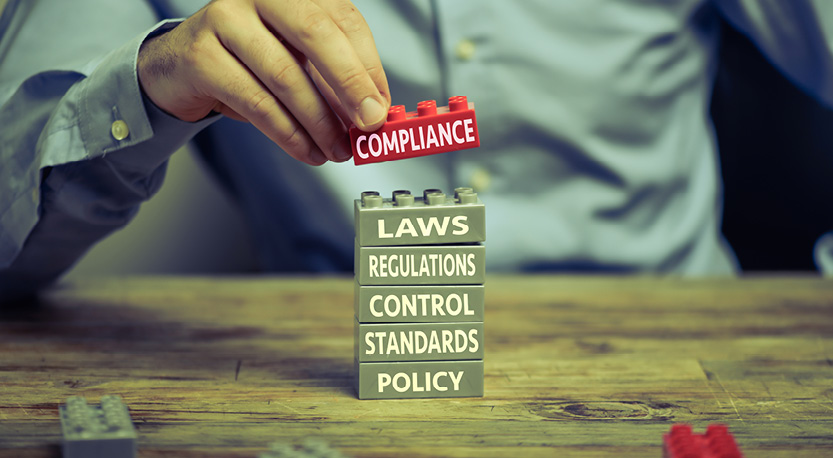 Man stacking a red plastic brick labeled Compliance onto a stack of gray plastic bricks labeled: Laws, Regulations, Control, Standards, and Policy