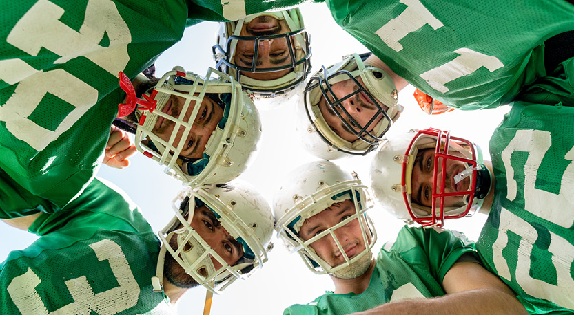 6 football players in helmets and green jerseys stand in a huddle looking down