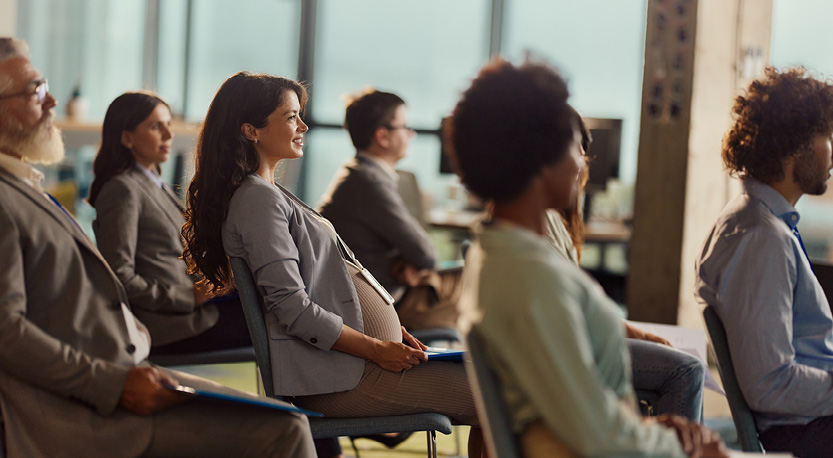 a group of business people sit in a chairs listening to a lecture, focus emphasized on the pregnant audience member