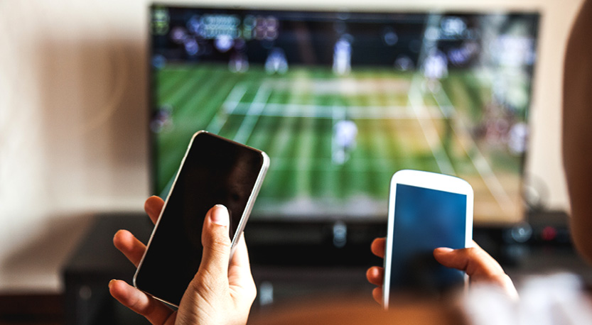 2 cell phones being held in anticipation in front of a tv screen showing a soccer game.