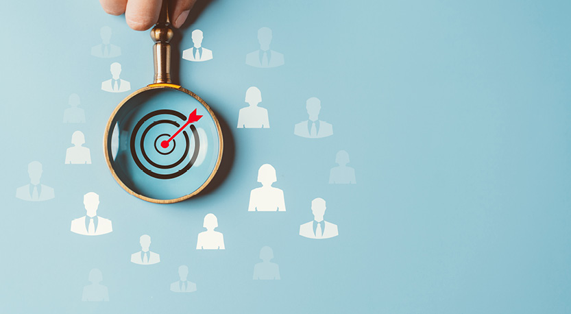 hand holding a magnifying glass over a target with an arrow in it surrounded by people icons.