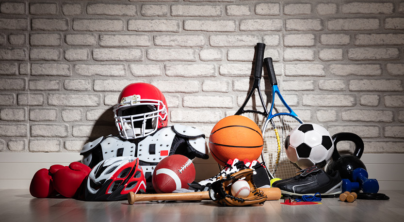 a pile of sports equipment in front of a brick wall (i.e. soccer ball, football, basketball, tennis ball, baseball bat, etc.)