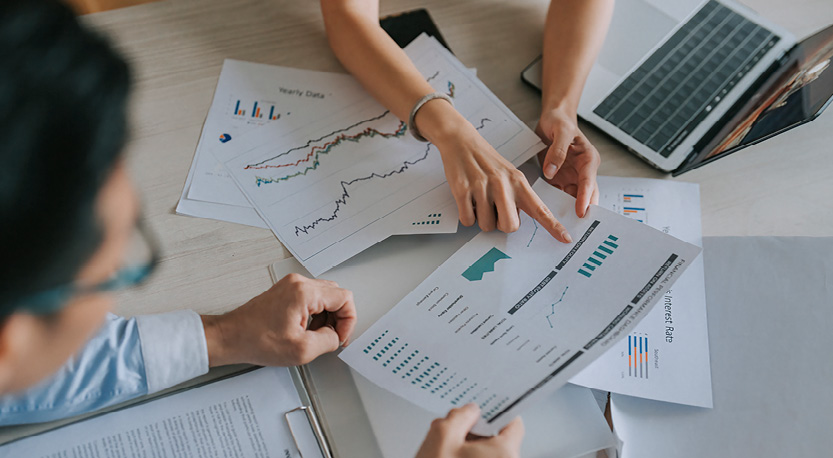 two people sit and point at printed charts spread across a desk.