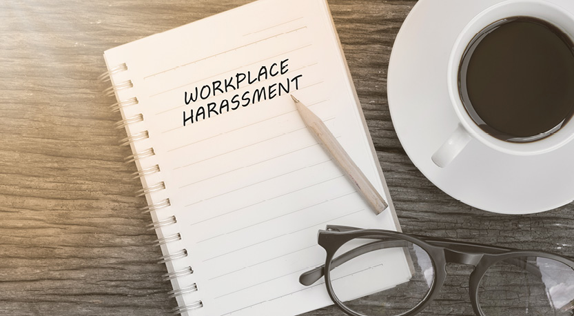 a notepad with a pencil resting on top reads 'Workplace harassment', surrounded by a coffee cup and pair of glasses/
