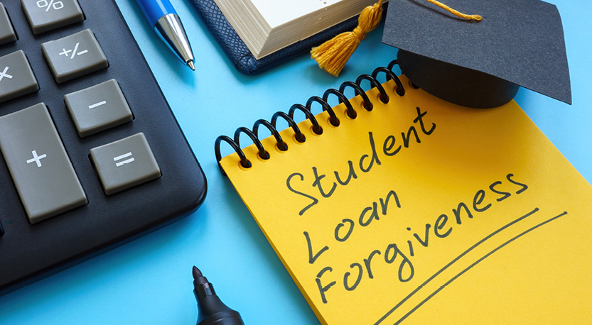 A calculator, notebook, pen and marker sit near a notepad that reads Student Loan Forgiveness, with a graduation hat placed on top.
